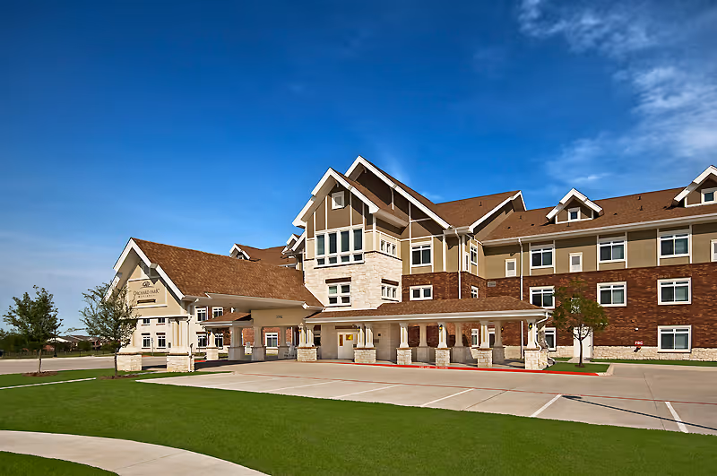Exterior view of a large senior living facility building with a covered entrance, multiple windows, and a well-maintained lawn under a clear blue sky.