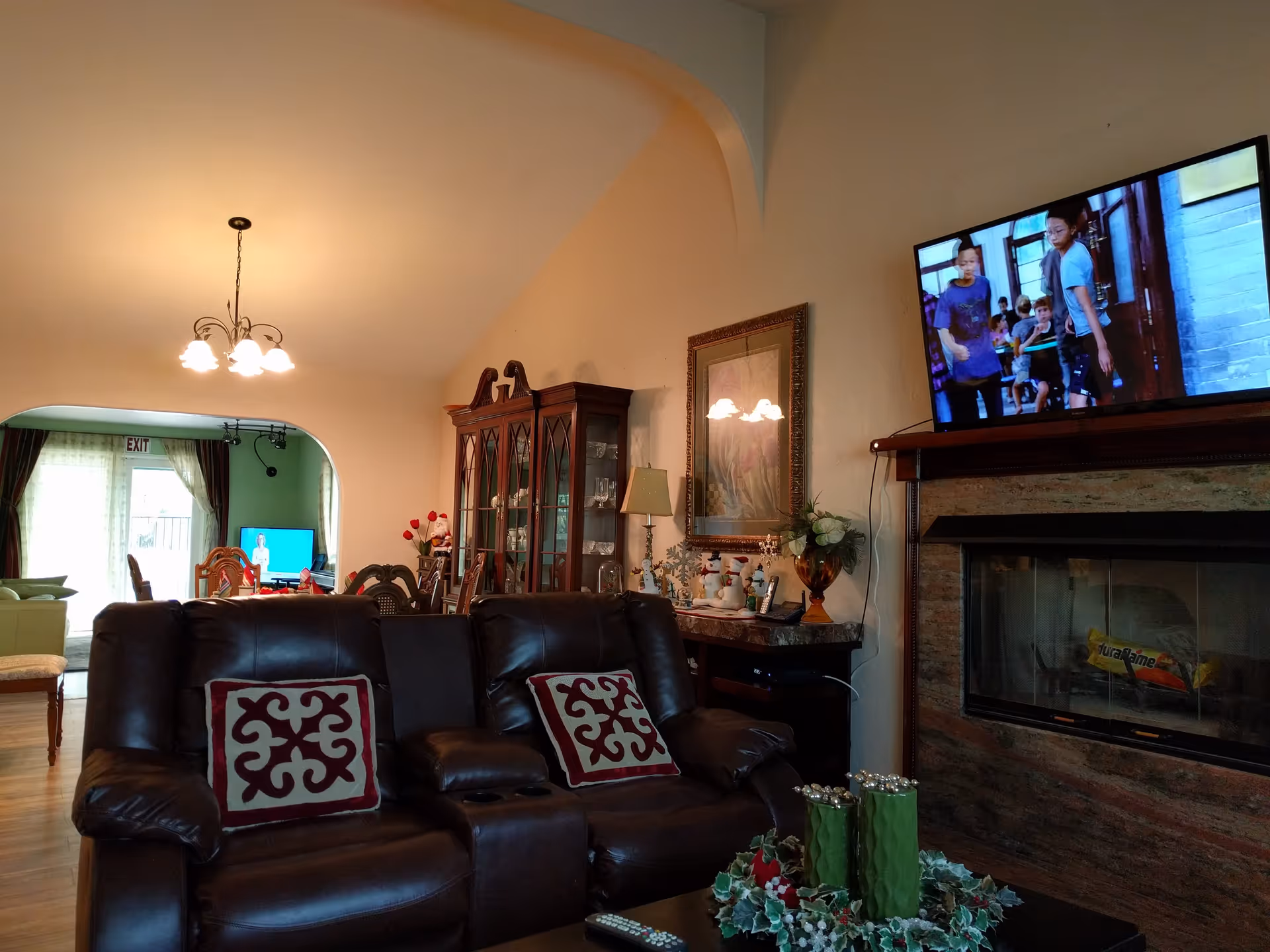 Interior view of a living room with two dark brown leather recliners featuring decorative pillows. A flat-screen TV is mounted above a fireplace on the right wall. A wooden cabinet with glass doors and a dining area with a chandelier are visible in the background. The room is decorated with holiday-themed items and has warm lighting.