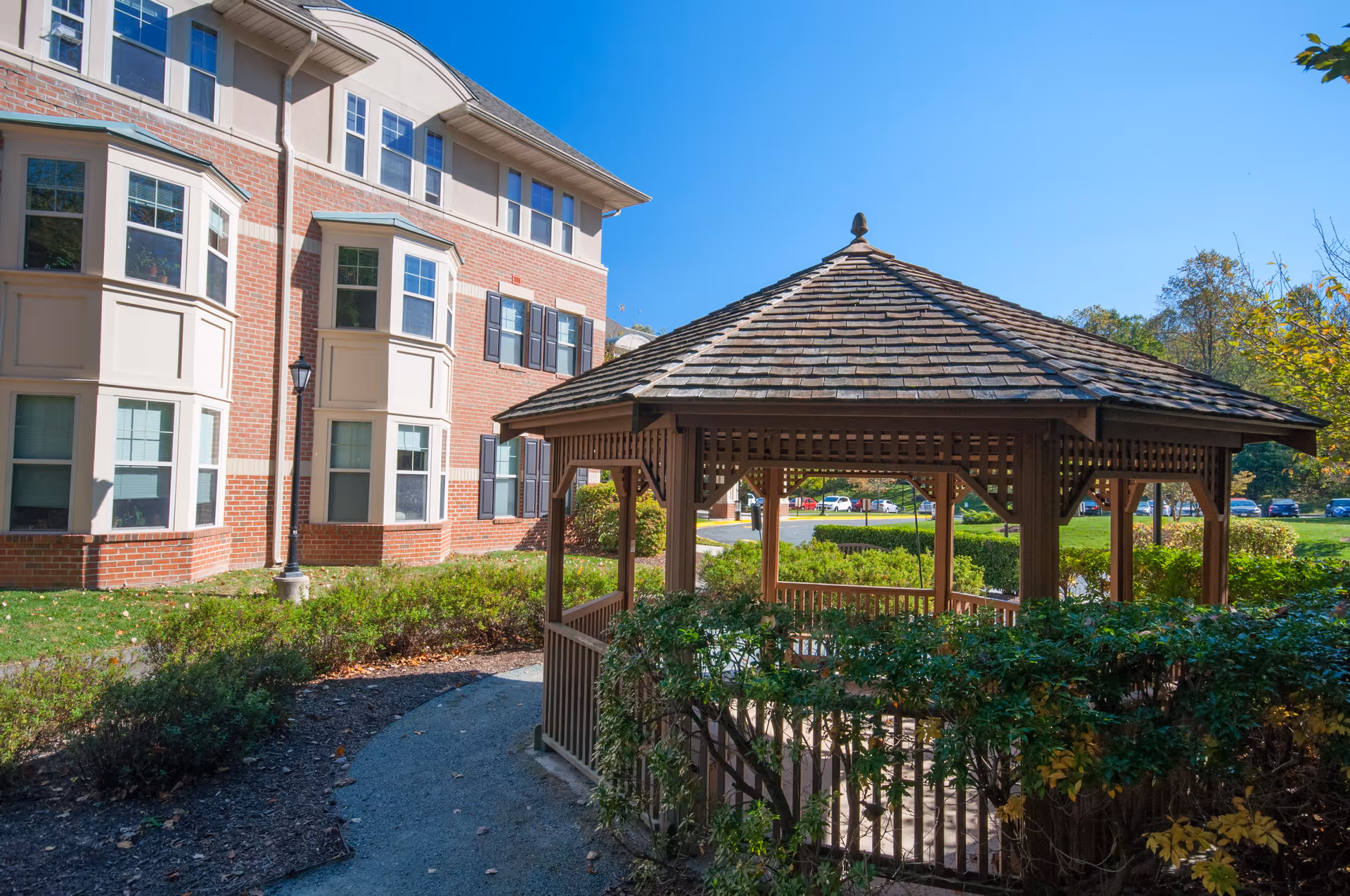 A wooden gazebo surrounded by green bushes and a paved pathway, located outside near a multi-story brick building with multiple windows under a clear blue sky.