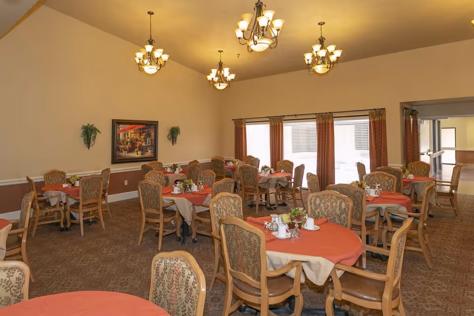 A dining room with multiple round tables covered with red and beige tablecloths. Each table is set with cups, glasses, and small flower arrangements. The room has patterned carpet, beige walls with a brown lower trim, several chandeliers hanging from the ceiling, and large windows with curtains letting in natural light.