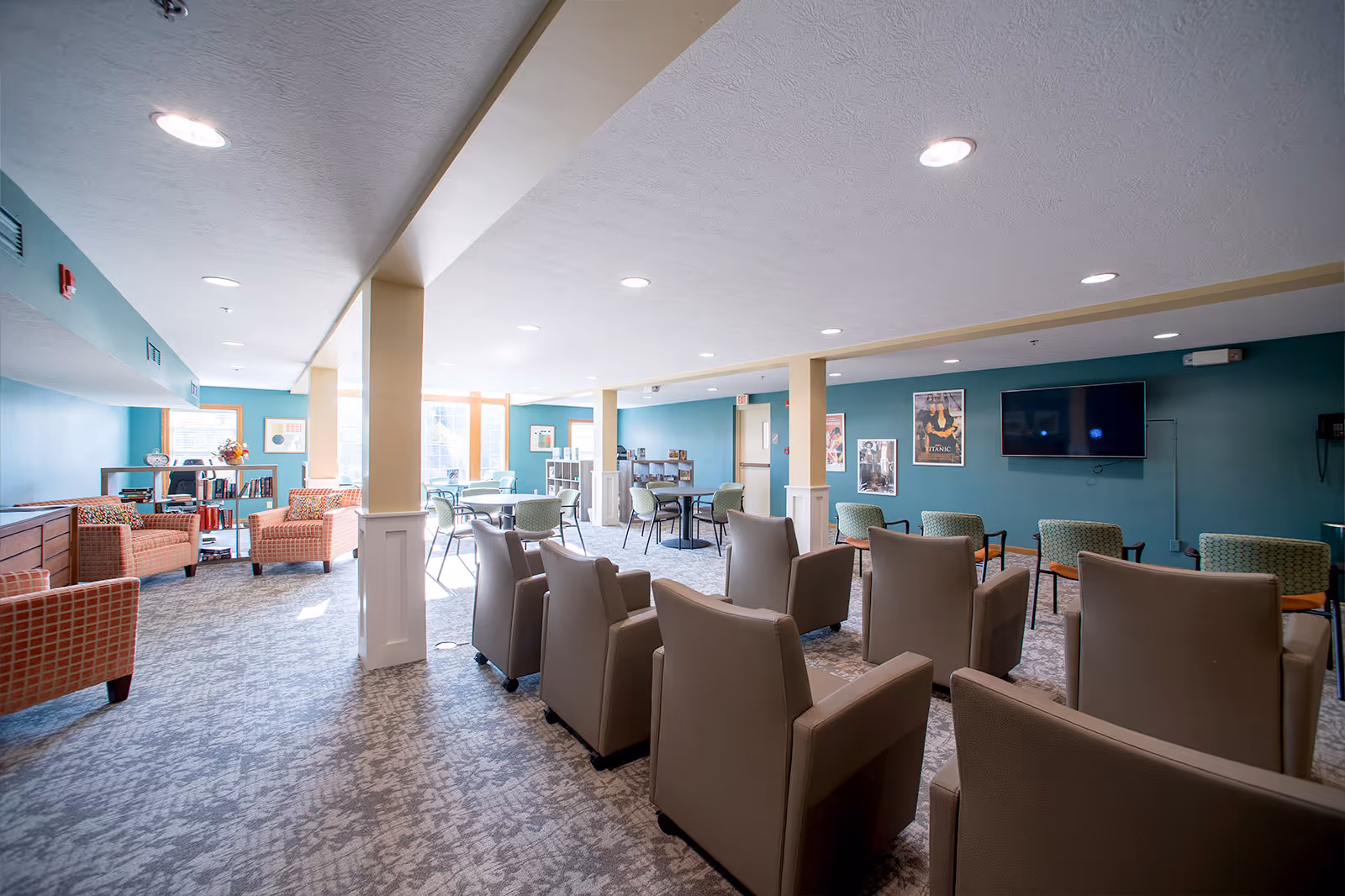 Spacious senior living common room with rows of armchairs facing a wall-mounted TV, plus tables and additional seating near windows.
