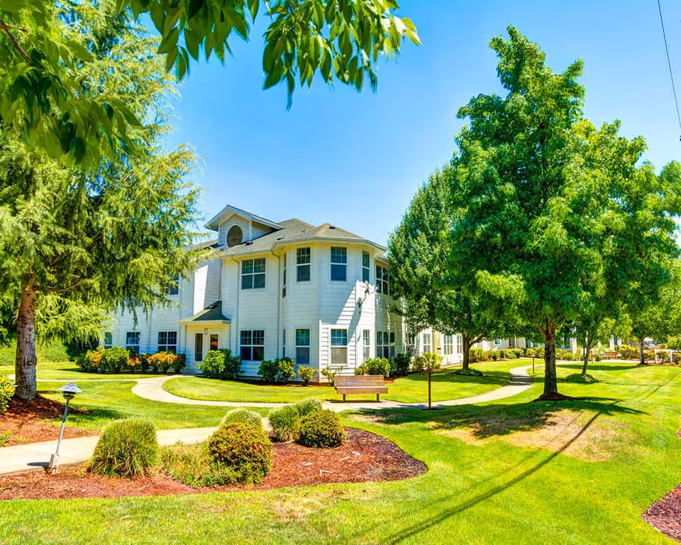 A bright and sunny outdoor view of a white two-story retirement and assisted living building surrounded by well-maintained green lawns, trees, bushes, and walking paths with a wooden bench.