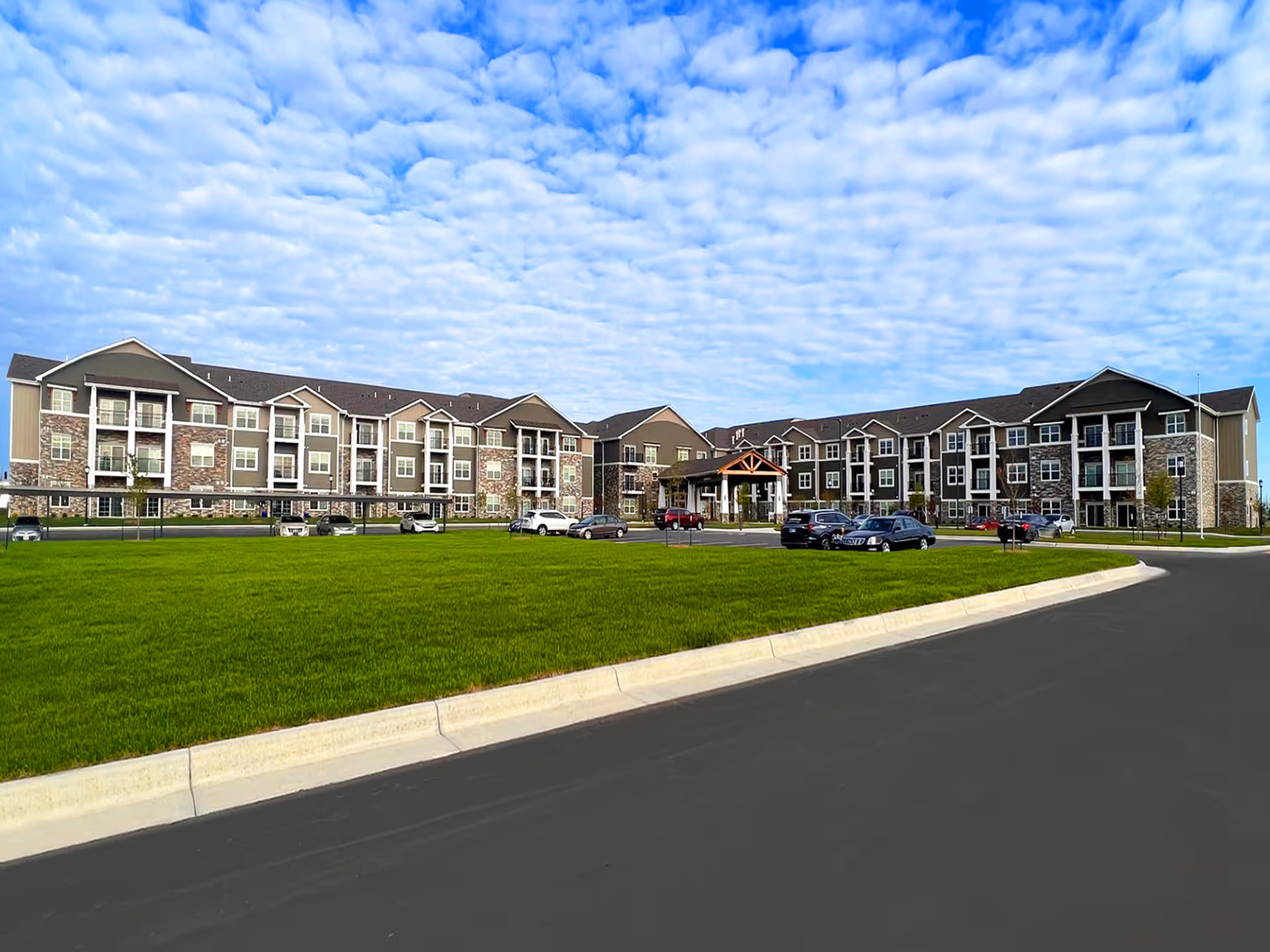 Wide exterior view of The Rushwood Senior Living facility showing a large three-story building with a combination of stone and siding facade, multiple balconies, a covered entrance, a green lawn in the foreground, several parked cars, and a partly cloudy sky above.