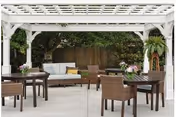 Outdoor seating area with a white pergola overhead, featuring a cushioned sofa, several wicker chairs, and wooden tables with flower arrangements, surrounded by greenery and a wooden fence.