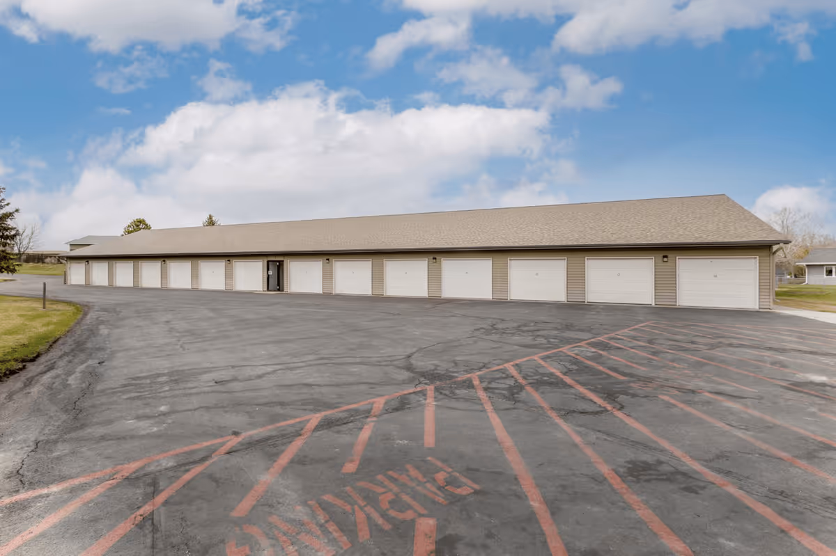 A long single-story building with multiple white garage doors, situated in a paved parking area with red painted lines and the word 'PARKING' visible on the asphalt. The sky is partly cloudy and there is some grass visible on the left side.
