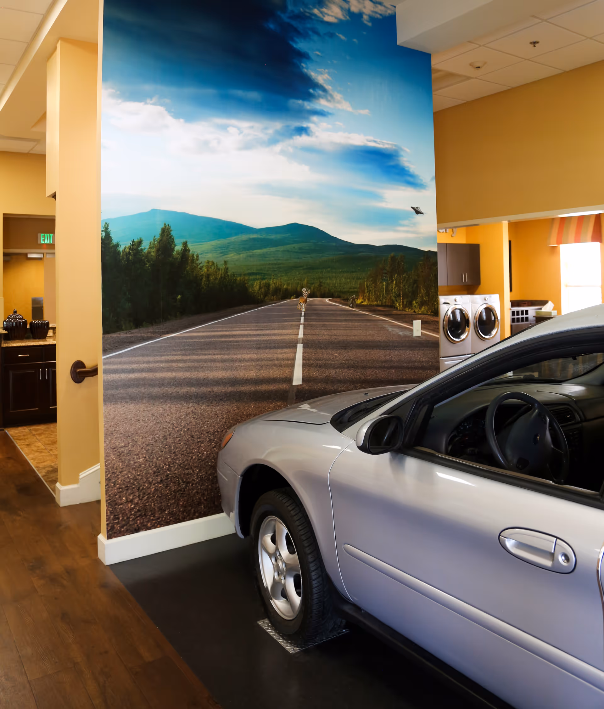 Indoor scene showing a silver car parked on a black floor mat next to a wall with a large mural of a road leading into a forested mountain landscape under a partly cloudy sky. The room has yellow walls, wooden flooring, and a laundry area with a washer and dryer visible in the background.