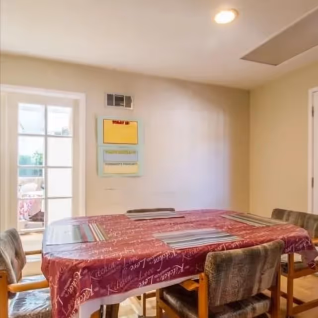A dining room with a rectangular table covered with a red tablecloth featuring white script. Four cushioned chairs with wooden armrests surround the table. On the wall, there is a small bulletin board with some notices. A glass door on the left side leads to an outdoor area.
