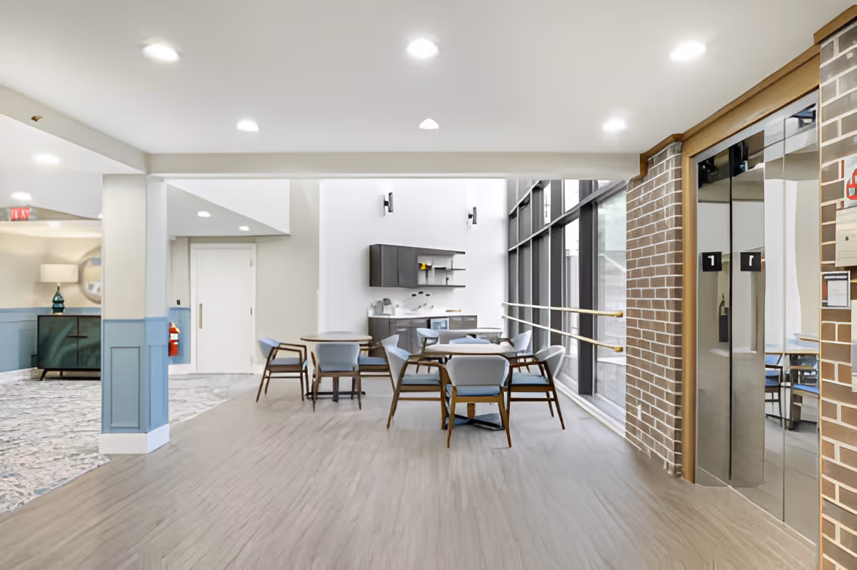 Bright communal seating area with tables and chairs, large floor-to-ceiling windows and elevators along a brick wall.