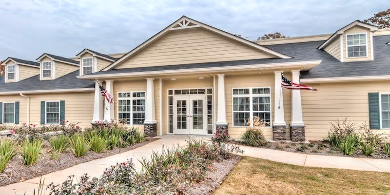 Front exterior view of a single-story building with beige siding, white columns, and multiple windows. There are two American flags on either side of the entrance, a walkway leading to the double glass doors, and landscaped bushes and plants in front of the building under a cloudy sky.
