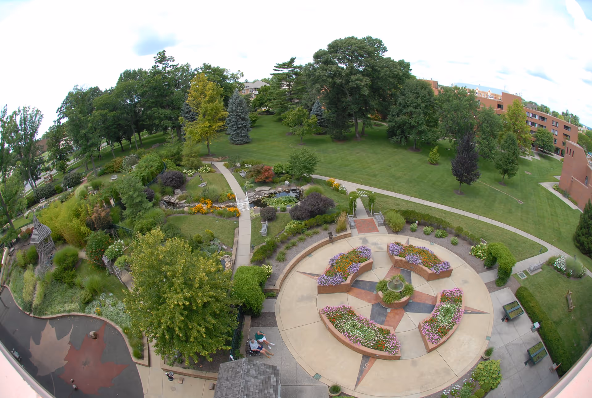 Aerial view of a landscaped garden area at Maple Knoll Village featuring a circular flower bed with colorful flowers arranged in sections around a central fountain. Surrounding the flower bed are paved walkways, benches, lush green grass, various trees, shrubs, and a small pond with rocks. Two people are sitting on a bench near the bottom of the image. Buildings are visible in the background.
