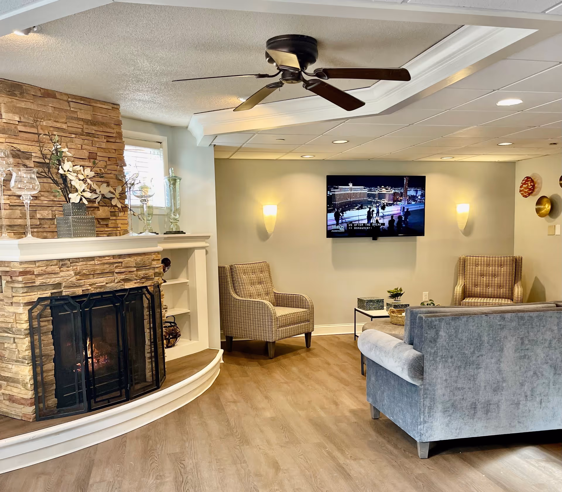 A cozy living room area with a stone fireplace on the left, a ceiling fan, two plaid armchairs, a gray sofa, a wall-mounted TV, and soft lighting on the walls. The floor is wood, and there are decorative items on the fireplace mantel and a small coffee table in front of the sofa.