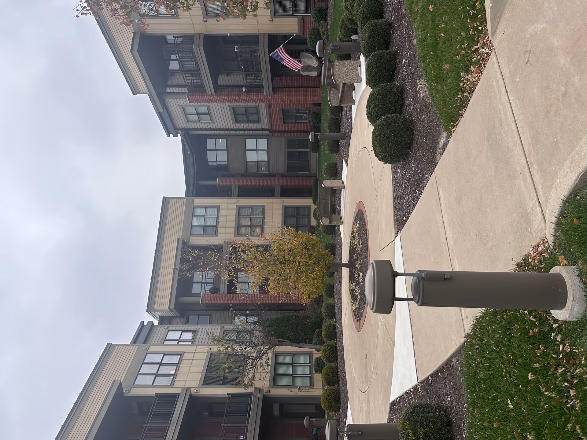 Outdoor courtyard area of a senior living facility with a circular paved walkway surrounding a small tree in the center, benches, trimmed bushes, and a three-story building with balconies and windows in the background under a cloudy sky.