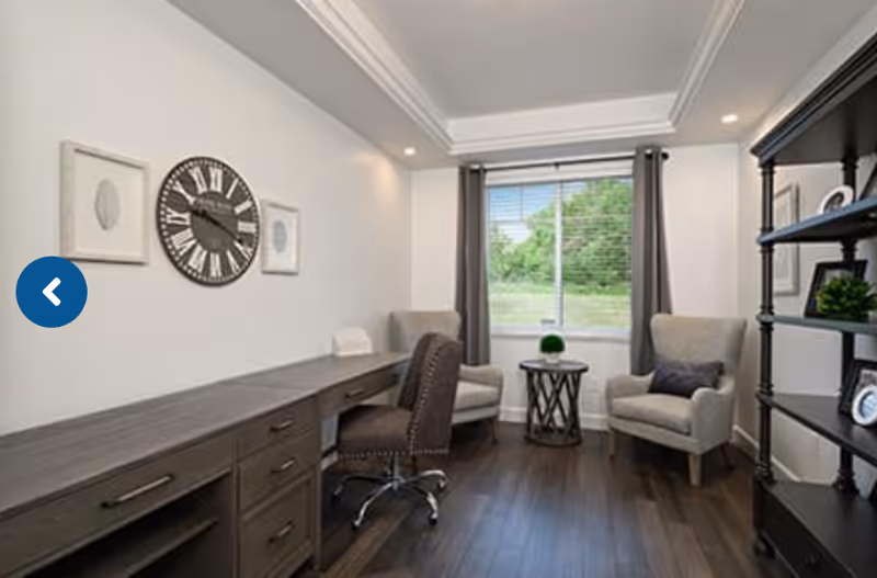 A well-lit interior room with a large window showing greenery outside. The room features a long wooden desk with a rolling chair, two cushioned armchairs with a small round table between them, and a tall black shelving unit with decorative items. The walls are white with crown molding and recessed lighting.
