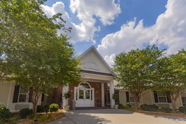 Front exterior view of Rock Hill Post Acute Care Center showing a brick building with white trim, a peaked roof entrance, large white double doors, and surrounded by green trees under a partly cloudy blue sky.