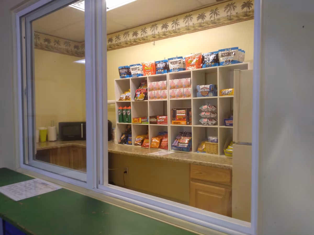 View through a window into a small snack room with shelves filled with various snacks including Hershey's, Reese's, Twix, and chips. There is a microwave on the countertop and a refrigerator to the right. The room has yellow walls with a palm tree border near the ceiling.