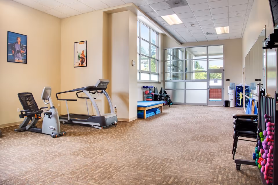 A spacious fitness room with exercise equipment including a recumbent bike and a treadmill. The room has large windows letting in natural light, posters on the wall, a bench with exercise mats underneath, and racks holding various weights and fitness accessories.