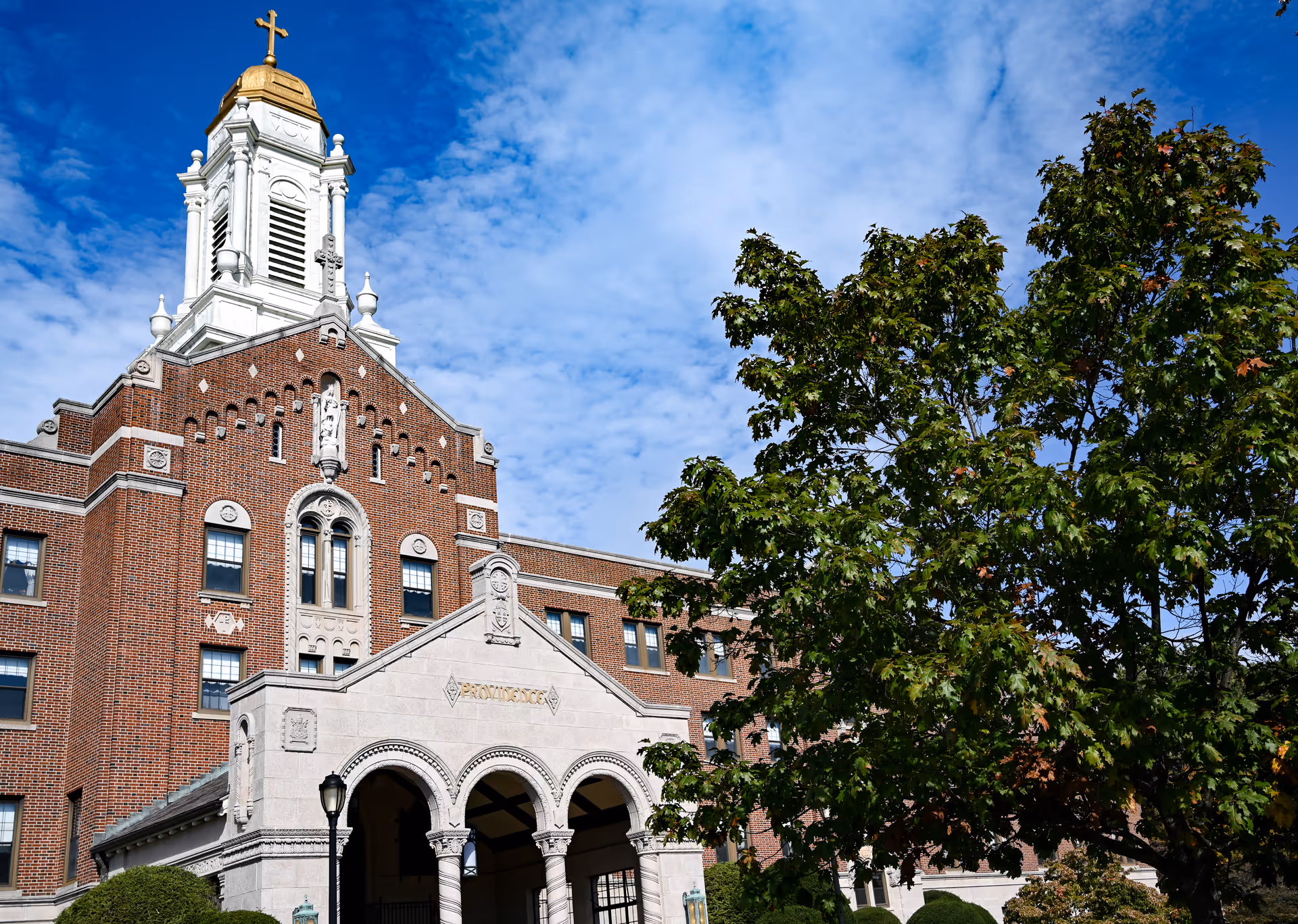 Exterior view of a large brick building with a white and gold cupola topped with a cross. The building has arched entranceways and decorative architectural details. A large tree with green leaves is visible on the right side under a partly cloudy blue sky.