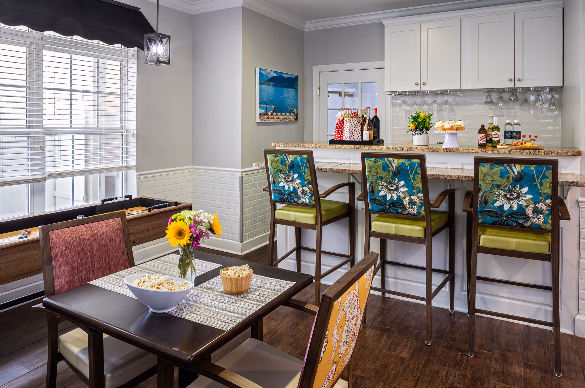 A bright and cozy dining area with a dark wooden table set for snacks including popcorn and a vase with sunflowers. Behind the table are three high chairs with floral-patterned backs at a kitchen counter. The kitchen counter has bottles of wine, cupcakes, and other beverages. A foosball table is visible near a window with white blinds, and a scenic wall picture hangs on the light gray wall.
