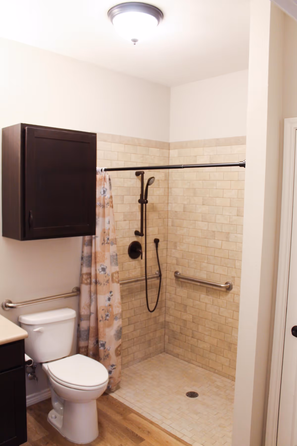 Accessible bathroom with a tiled walk-in shower, grab bars, handheld showerhead, shower curtain, toilet, and a dark wood wall cabinet.