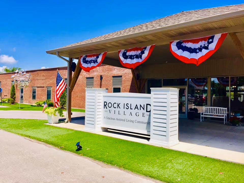 Entrance of Rock Island Village Assisted Living facility with a covered porch decorated with red, white, and blue bunting. An American flag is displayed near the entrance, and a white sign with the facility's name and tagline is prominently placed in front. The building is made of red brick and there is a bench visible under the porch.