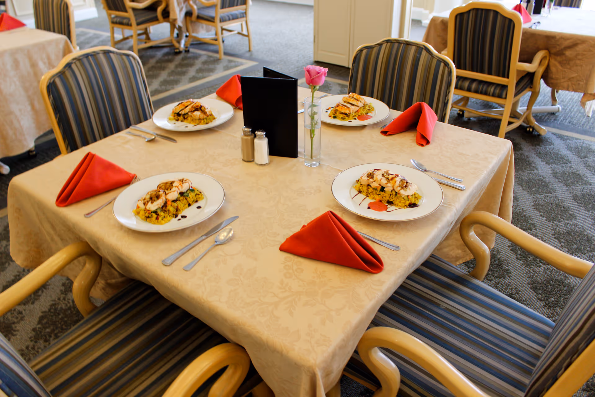 A dining table set for four with plates of food, red folded napkins, silverware, salt and pepper shakers, a black menu, and a single pink rose in a vase. The chairs around the table have striped upholstery, and other similarly set tables are visible in the background.