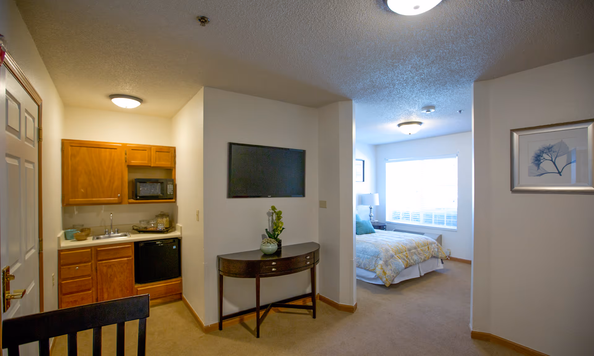 Interior view of a senior living facility apartment showing a small kitchenette with wooden cabinets, a microwave, sink, and mini fridge on the left. In the center, a wall-mounted flat-screen TV is above a small dark wooden console table with a decorative plant. To the right, a bedroom area is visible with a bed covered in a yellow and gray patterned comforter, a bedside table with a lamp, and a large window with blinds letting in natural light. The walls are painted white and the floor is carpeted.
