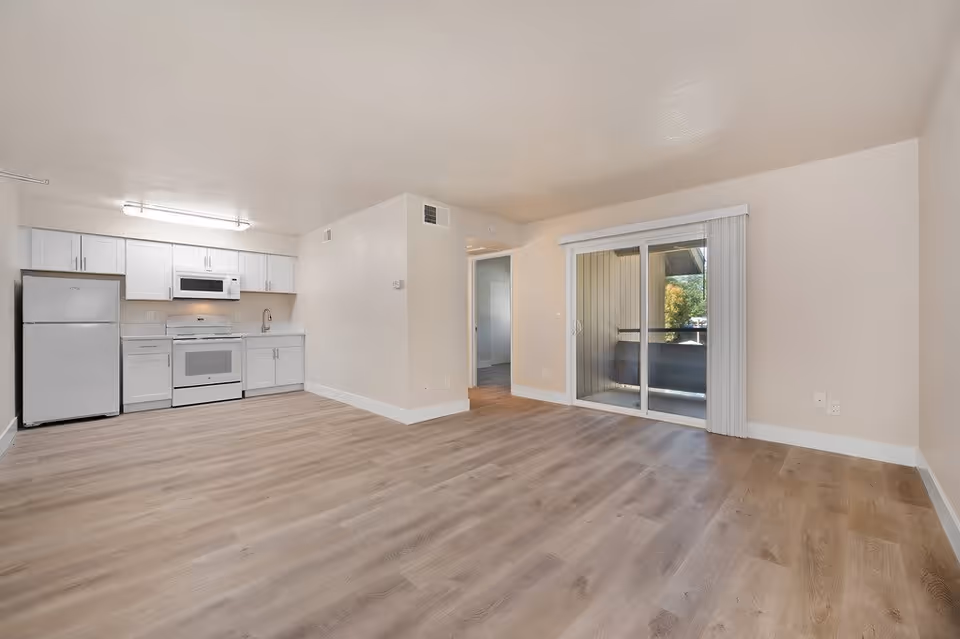 An empty apartment interior featuring a small kitchen area with white cabinets, a refrigerator, stove, and microwave on the left side. The room has light wood flooring and beige walls. There is a sliding glass door with vertical blinds leading to a small balcony or outdoor space. A doorway leads to another room in the background.
