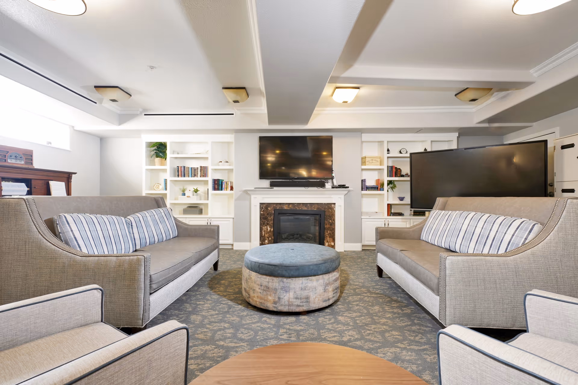 A cozy living room with two gray sofas facing each other, each with striped cushions. A round ottoman is centered between the sofas on a patterned carpet. In the background, there is a fireplace with a mounted flat-screen TV above it, flanked by white built-in bookshelves filled with books and decorative items. The room has a bright and clean atmosphere with soft lighting.