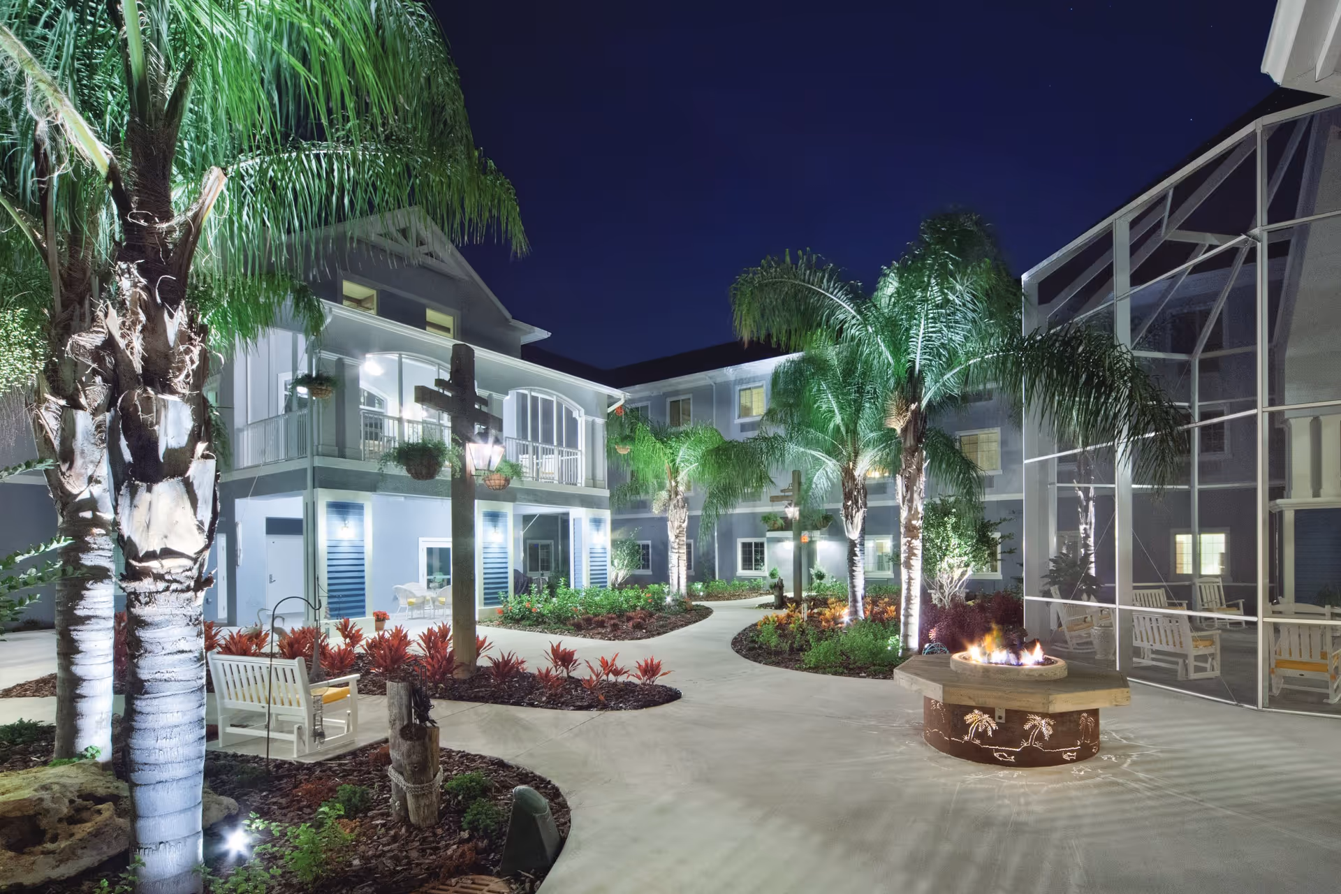 Night view of a landscaped courtyard with palm trees, walkways, balconies and a lit fire pit.