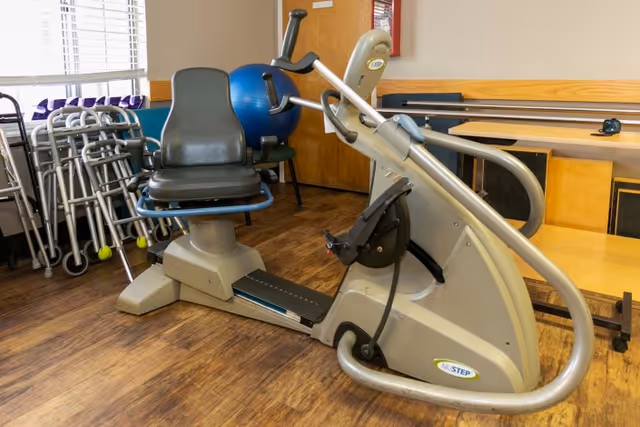 A recumbent exercise bike in a room with wooden flooring, several walkers lined up against the wall, a blue exercise ball, and a wooden table with chairs in the background.