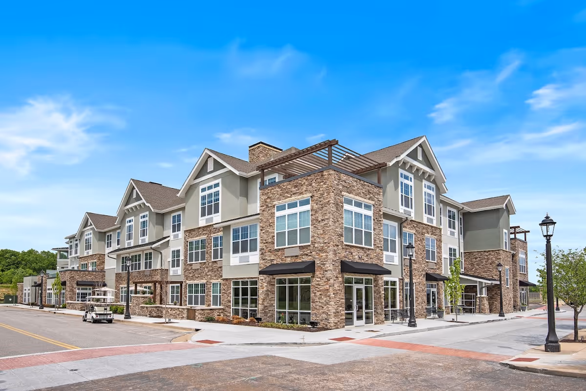 Exterior view of a modern three-story senior living facility building with stone and beige siding, large windows, and a clear blue sky in the background. The building is situated on a paved street with sidewalks, street lamps, and some small trees.
