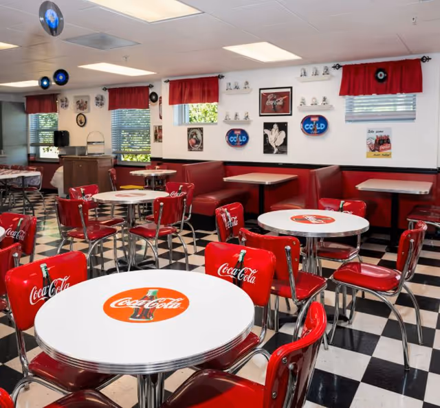A retro-style dining room with black and white checkered floor, red Coca-Cola branded chairs, and white round tables featuring Coca-Cola logos. The walls are decorated with vintage Coca-Cola signs and memorabilia. Red curtains hang over the windows, and there are red cushioned booth seats along the walls.