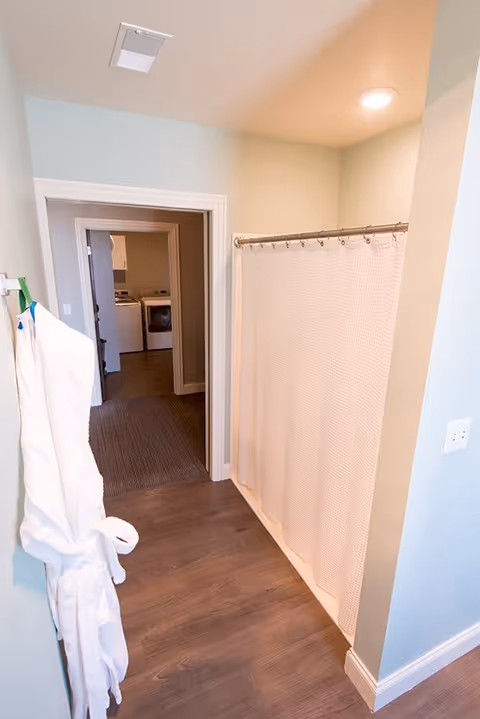 Interior view of a bathroom area with a white shower curtain on a rod to the right and two white bathrobes hanging on hooks on the left wall. The floor is wood-style laminate, and there is a doorway leading to another room with a washer and dryer visible in the background.