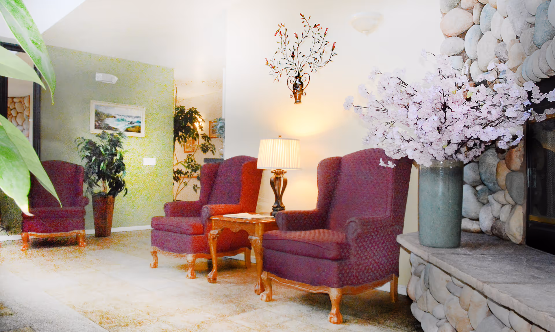Cozy lounge area with three burgundy armchairs, a side table with a lamp, potted plants, and a vase of pink blossoms by a stone fireplace.