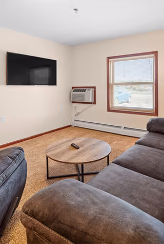 A cozy living room with a brown carpet, a large dark gray sectional sofa, a round wooden coffee table with a remote control on it, a wall-mounted flat screen TV, a window with blinds, and a wall-mounted air conditioning unit below the window.