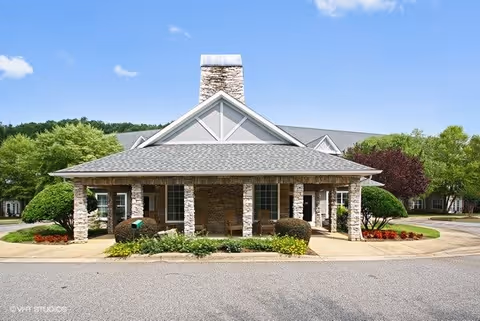Front exterior view of a single-story building with a stone facade, a covered porch supported by stone pillars, and a central chimney. The building is surrounded by well-maintained landscaping including bushes, flowers, and trees under a clear blue sky.