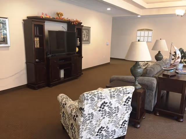 Carpeted lounge area with a TV entertainment center, patterned armchair, sofa, lamps, and side tables.