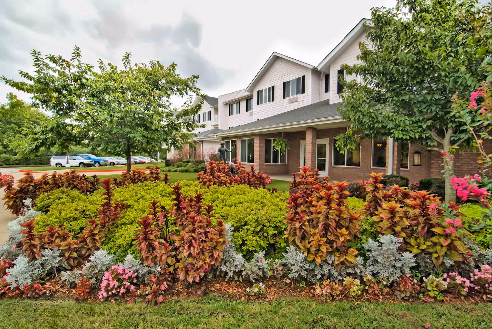 Front exterior of a two-story senior living building with landscaped flowerbeds, trees, and a driveway.