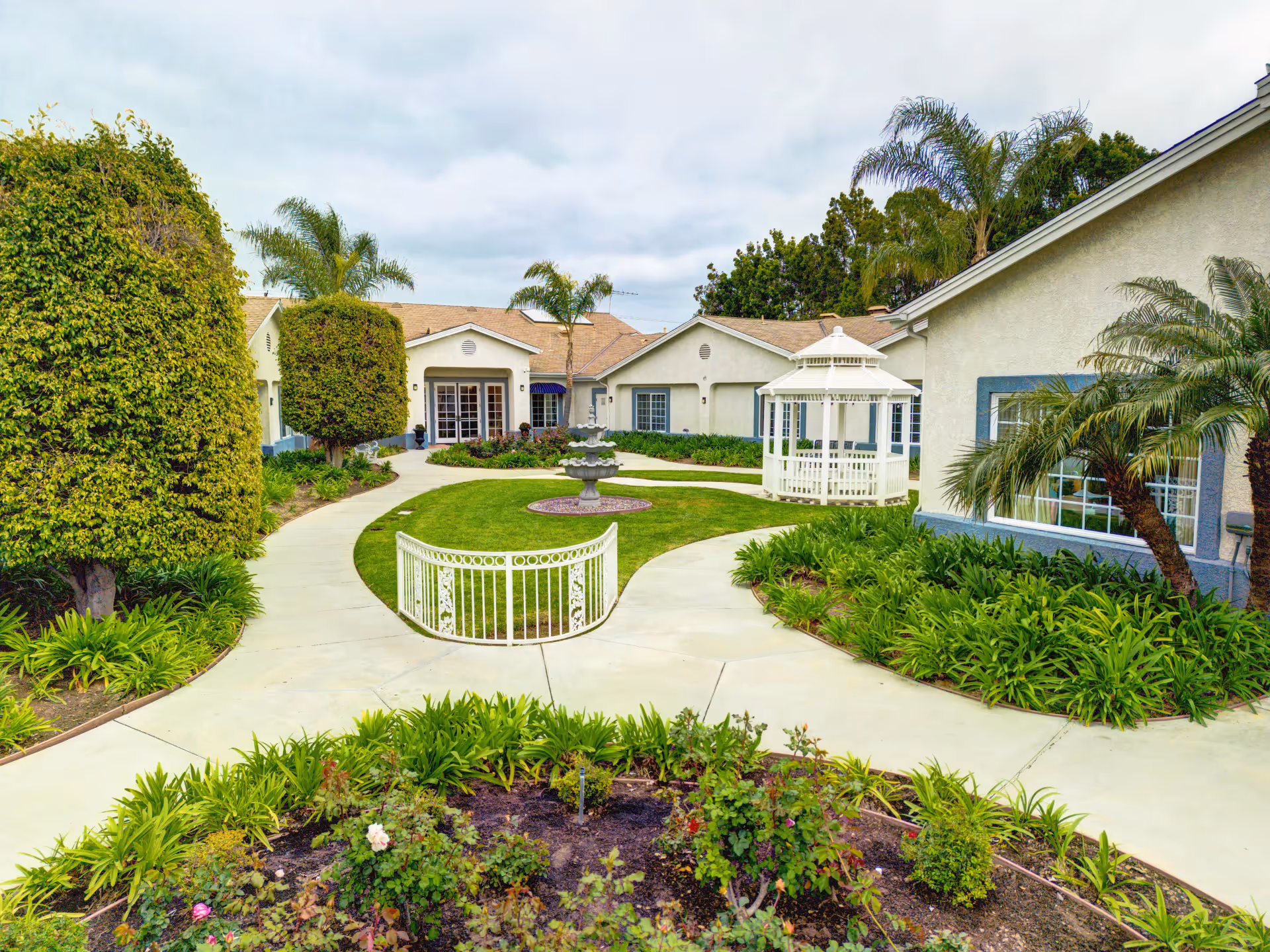 A landscaped outdoor courtyard at Ivy Terrace at Garden Grove Memory Care featuring a curved concrete walkway, trimmed bushes, palm trees, a white gazebo, and a multi-tiered fountain in the center of a grassy area, surrounded by single-story buildings.