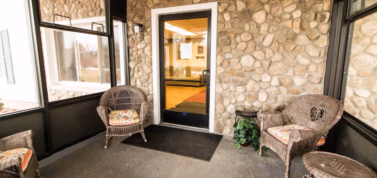 A cozy outdoor seating area with three wicker chairs featuring floral cushions, a small round wicker table, and a small plant on a stand. The area is enclosed with stone walls and large windows, and a glass door leads inside to a well-lit interior space.