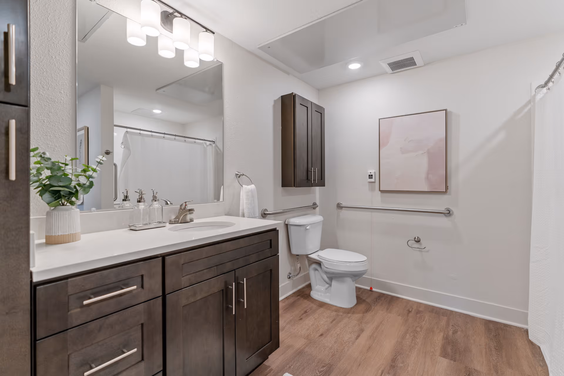 A clean and modern bathroom featuring a white toilet with grab bars on the walls, a dark wood vanity with a white countertop and sink, a large mirror above the sink, a small potted plant on the countertop, a towel ring with a white towel, a shower area with a white curtain, and a framed abstract artwork on the wall.