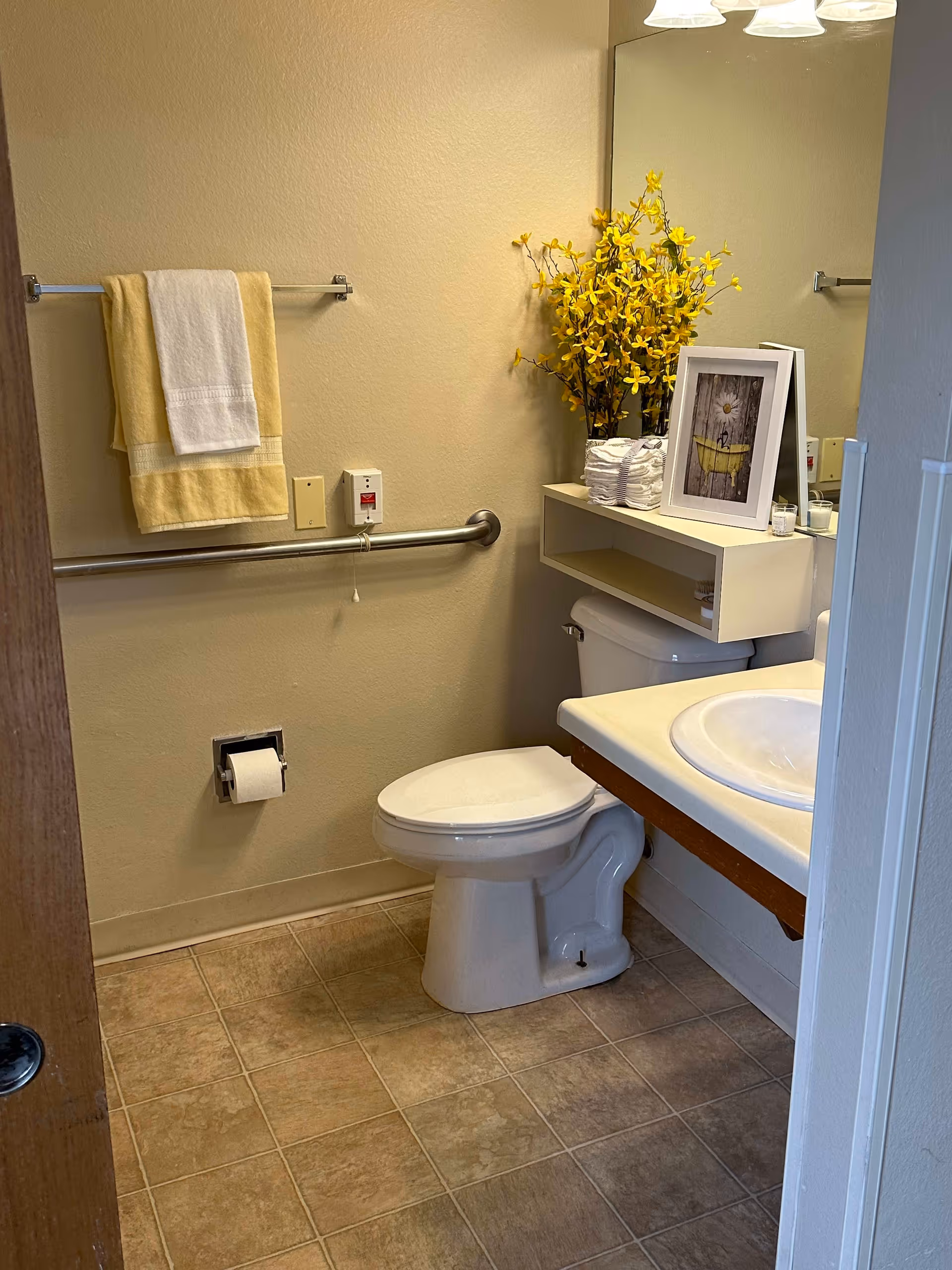 A clean bathroom with a white toilet, a sink with a countertop, and a large mirror above the sink. There is a towel rack on the wall with a yellow and a white towel hanging. A grab bar is installed on the wall next to the toilet. On top of the toilet tank, there is a shelf holding a framed picture, a stack of folded white towels, a small candle, and a vase with yellow flowers. The floor is tiled in a beige color.