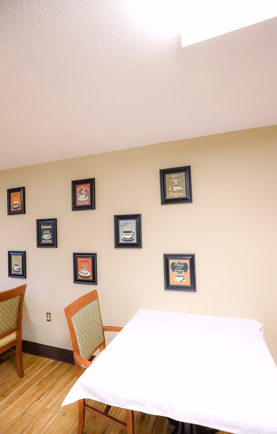 A small dining area with a table covered by a white tablecloth and two wooden chairs with green cushioned backs. The beige wall behind the table is decorated with eight framed pictures featuring coffee-themed artwork and text. The floor is wooden, and the ceiling has a bright light source.