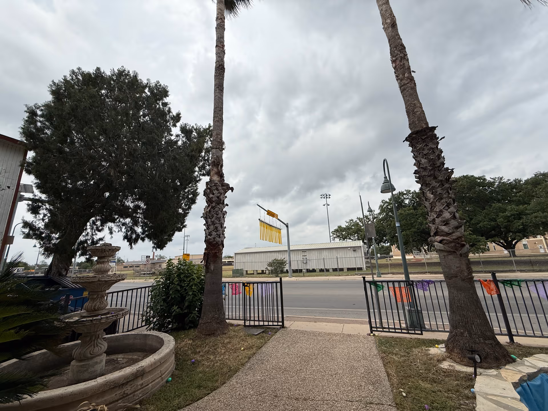 View from a gated entrance showing a walkway between two palm trees, a stone fountain, a street and buildings under a cloudy sky.
