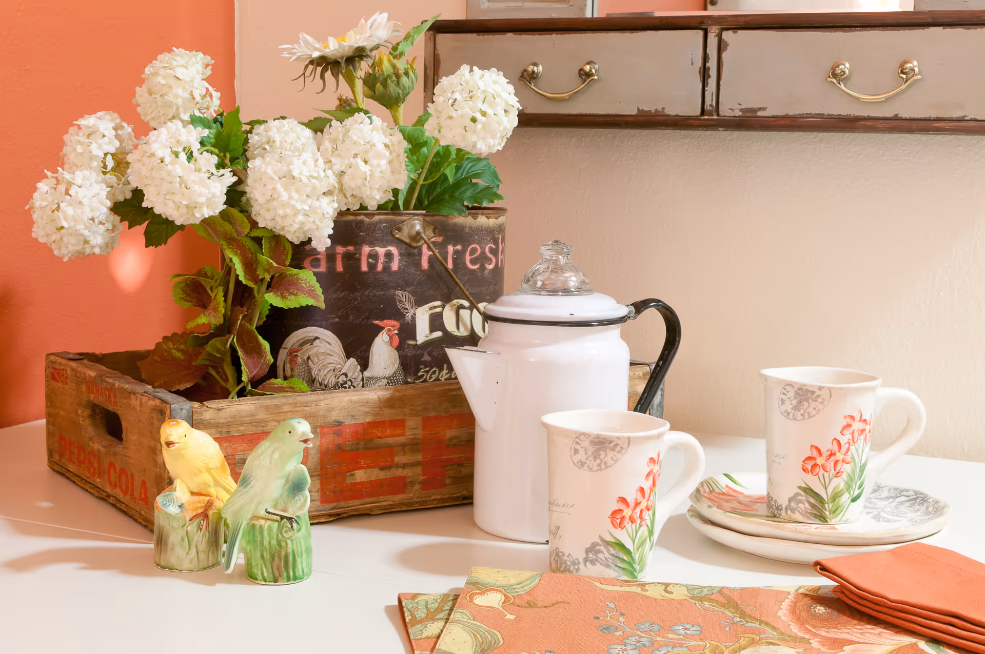 A decorative arrangement on a white table featuring a wooden crate with white flowers and green leaves, a white enamel coffee pot with a glass lid, two floral-patterned mugs on matching plates, two ceramic bird figurines, and a folded orange napkin with a floral placemat underneath. A wooden drawer with brass handles is mounted on the wall in the background.