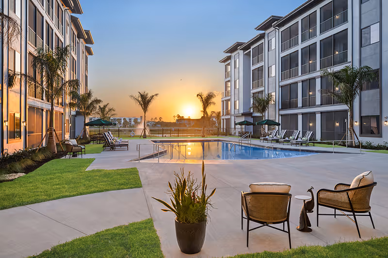 Outdoor swimming pool area at sunset with lounge chairs, umbrellas, palm trees, and two cushioned chairs with a small table on a concrete patio, surrounded by a multi-story residential building.
