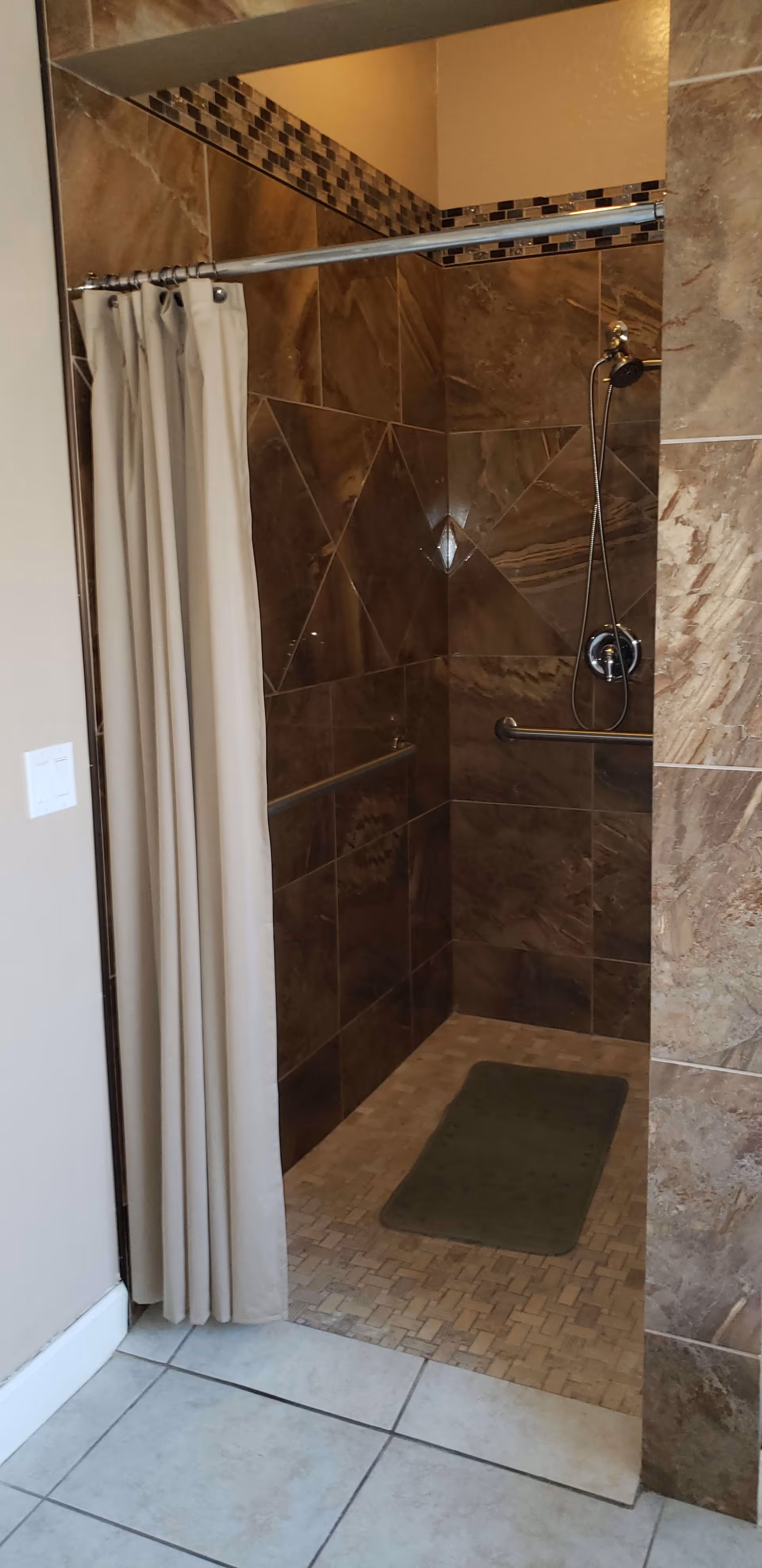 A tiled walk-in shower with brown and beige tiles, a silver showerhead, a grab bar, and a beige shower curtain partially drawn to the side. There is a green bath mat on the shower floor and light-colored tiles outside the shower area.