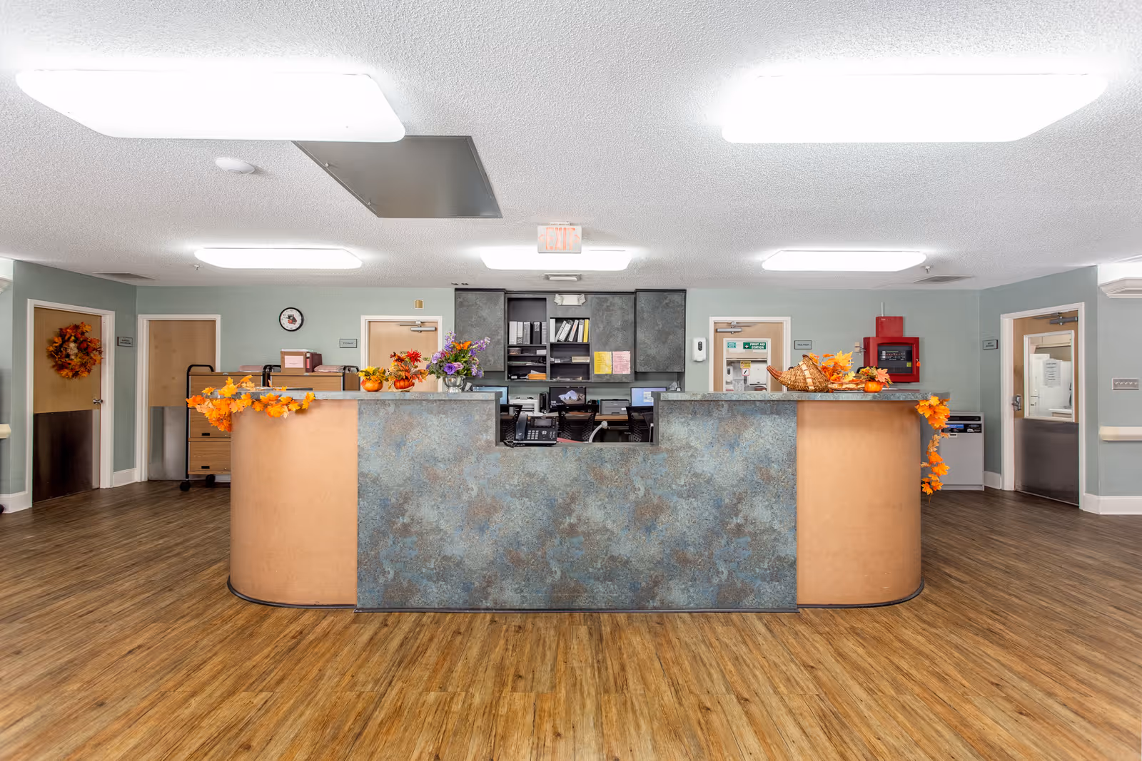 Curved reception desk in a nursing facility lobby decorated with autumn garlands and flowers, with wood flooring and doors in the background.
