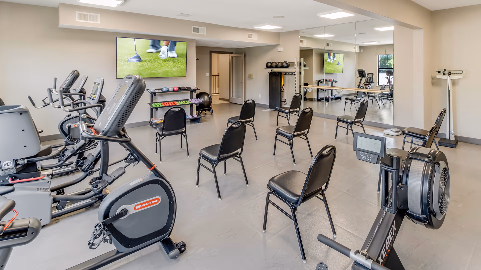 Interior fitness room with exercise machines, chairs arranged for group exercise, free weights, and a wall-mounted TV.
