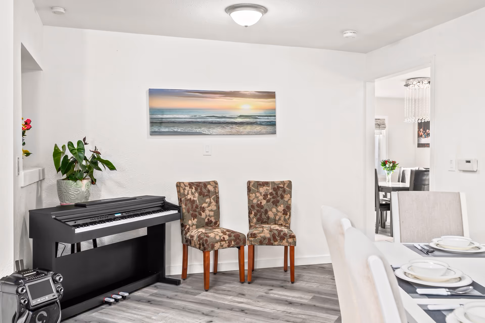 A bright interior room with a black electronic keyboard against a white wall, two floral-patterned chairs next to it, and a potted plant on top of the keyboard. A framed picture of a sunset over the ocean hangs on the wall above the chairs. To the right, part of a dining table set with white dishes and beige chairs is visible, and through an open doorway, another dining area with a chandelier and a vase of flowers on the table can be seen.